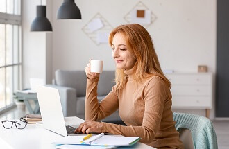 אישה שותה קפה מול מחשב ולידה דפים ועט