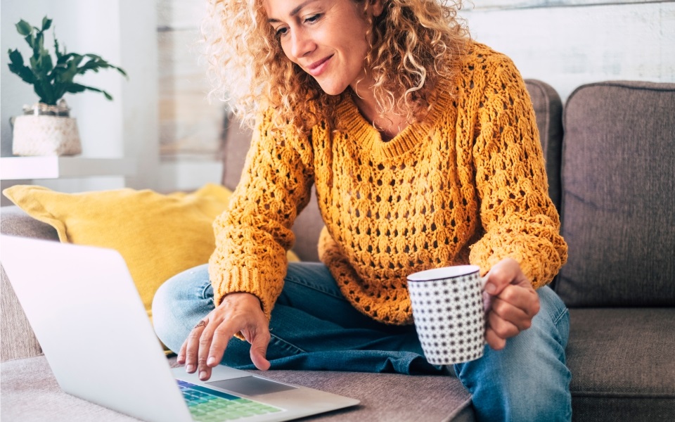 A woman with a mustard shirt and curls in front of a laptop is sitting on a couch