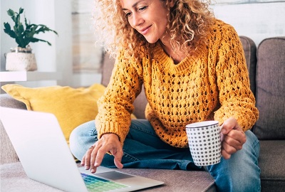 אישה עם חולצה בצבע חרדל מחזיקה ספל ביד אחת וביד השניה נוגעת בטאצ של מחשב נייד