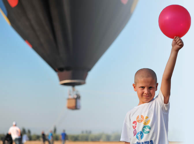 A boy in a shirt of larger-than-life holds a deep pink balloon and a hot air balloon behind him
